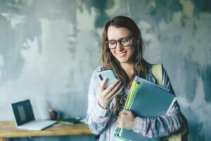 A positive hipster student on a college campus holds her textbooks and looks at her phone.